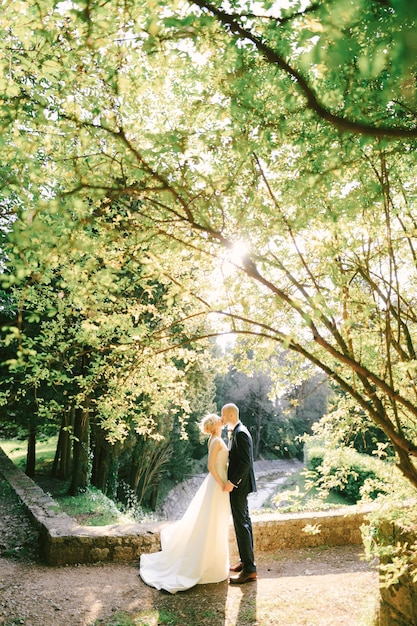 Groom in a black suit holds hands and kisses bride in a white dress standing on a path in a green