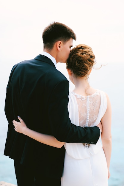 Groom in a black suit hugs and kisses bride in a white beautiful lace dress in the temple back view