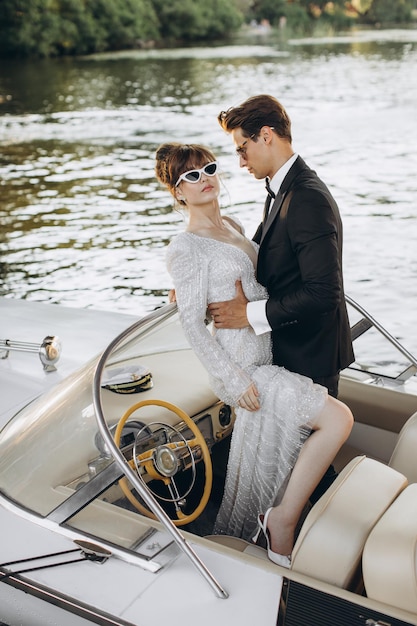 Groom in a black suit and sunglasses hugs bride in wedding dress on a yacht on a sunny day