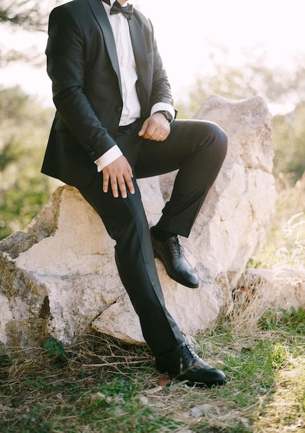 Groom in a black suit and white shirt sits on a stone in the park