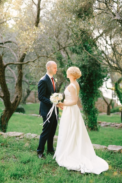 Groom in a blue suit stands in front of bride in a white dress in the park against the background of