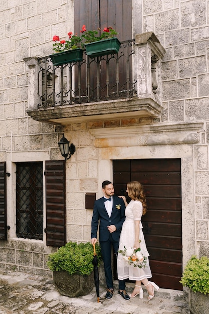 Groom and bride stand near the door of an old stone house with a balcony and shutters