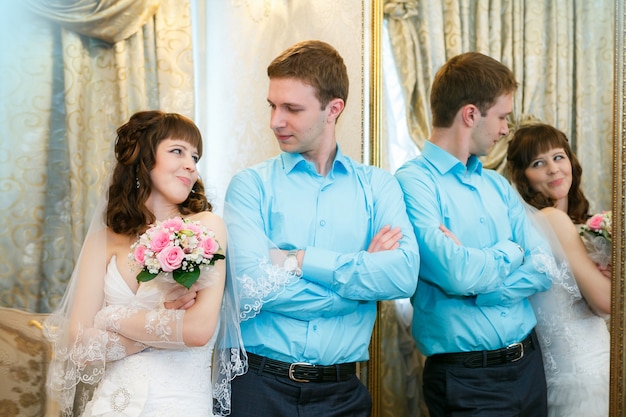 Groom and the bride stand near a mirror with a gold frame