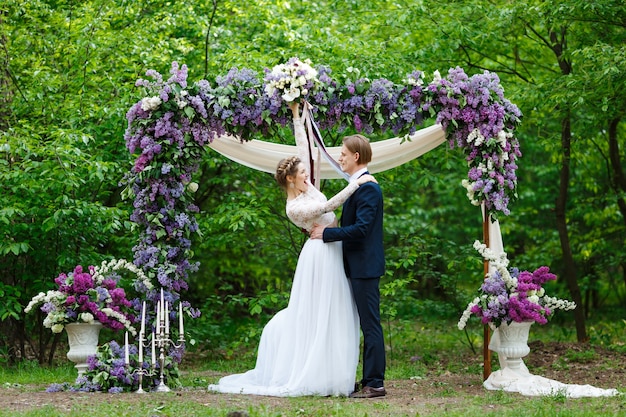 Groom and bride standing near arch with lilac flowers in vegetation background