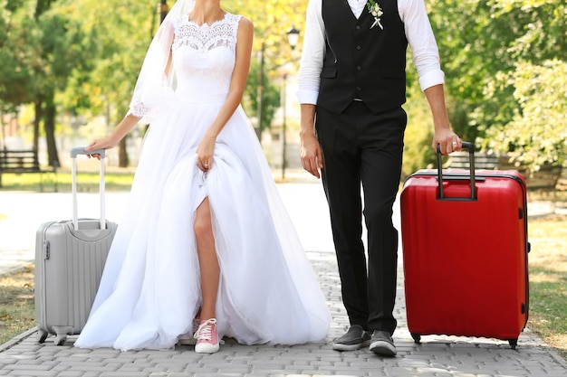 Groom and bride with suitcases walking in park