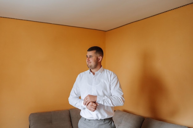 The groom buttons his shirt by the window man in a shirt Male portrait Photoshoot at the window Grooms collection Wedding day Handsome man groom buttoning his shirt in a hotel room