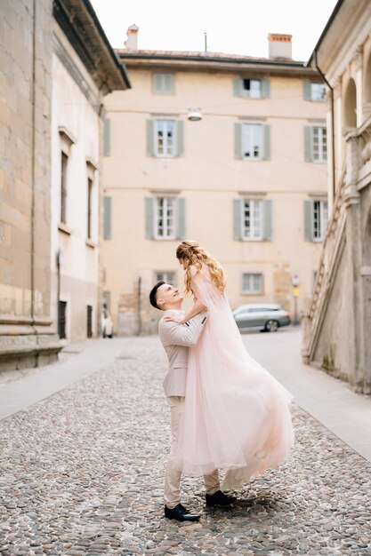 Groom circles bride in his arms standing on the paving stones near the old building in bergamo italy