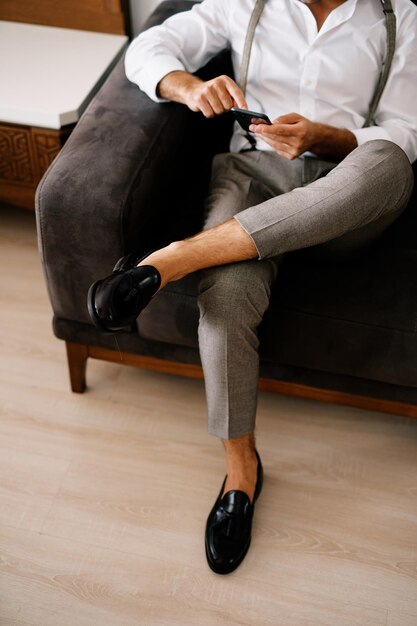 Groom clicks on a smartphone in his hands while sitting on the sofa cropped faceless