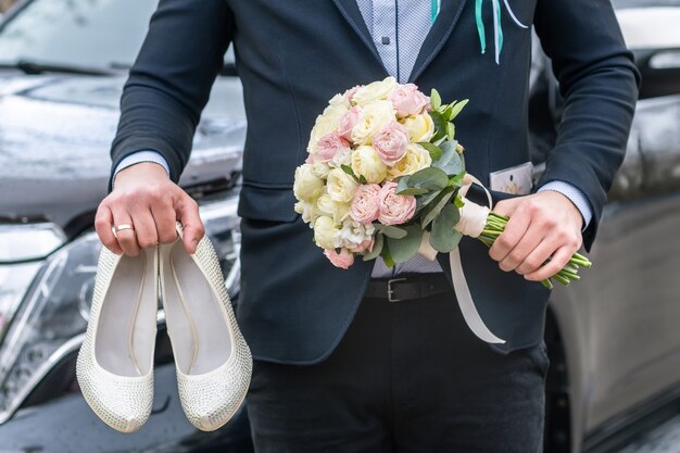 Groom dressed in blue suit holding a bridal bouquet of roses and white female wedding shoes on a car background.