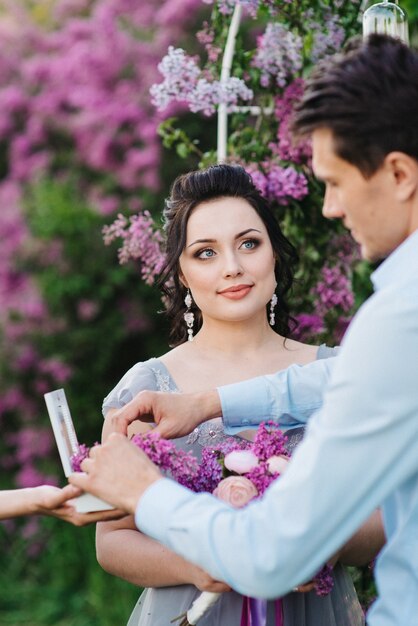 The groom dresses the bride with wedding rings near the lilac arch