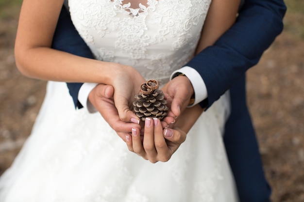 Groom embraces bride in a pine forest, their hands holding a lump