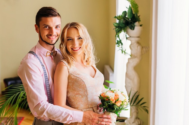 The groom embraces her bride who holds a wedding bouquet in a room with decorations