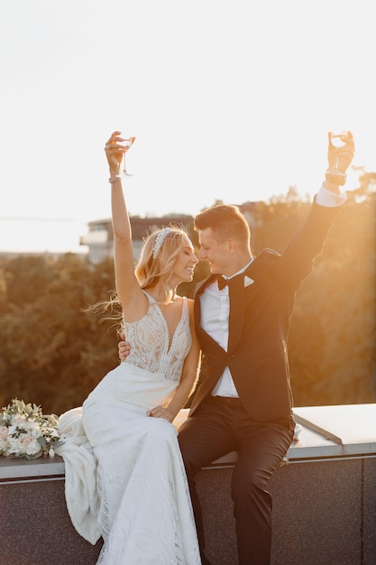 Photo groom in formal suit, woman in wedding dress raised hands high with glasses of wine or champagne. sitting on roof. against background of city,