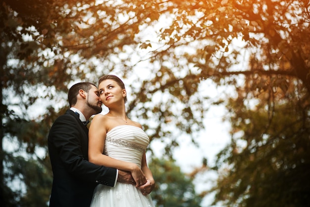 Lo sposo abbraccia dolcemente la sposa con un bouquet da dietro in un parco