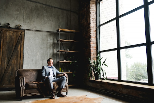 The groom in a in a gray suit, white shirt and a bow tie is sitting on a brown leather sofa.