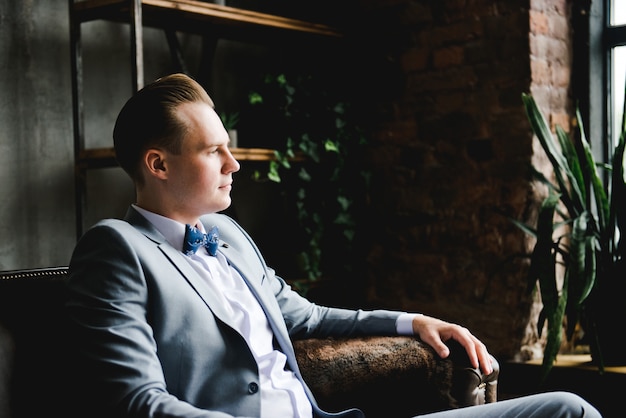 The groom in a in a gray suit, white shirt and a bow tie is sitting on a brown leather sofa.