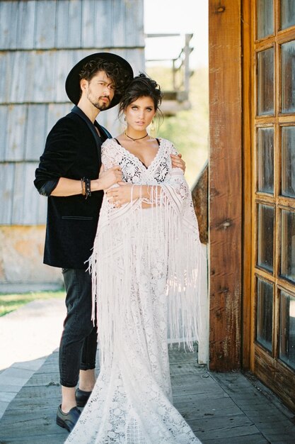 Groom in a hat holds his hands on the shoulders of bride standing on the threshold of a wooden house