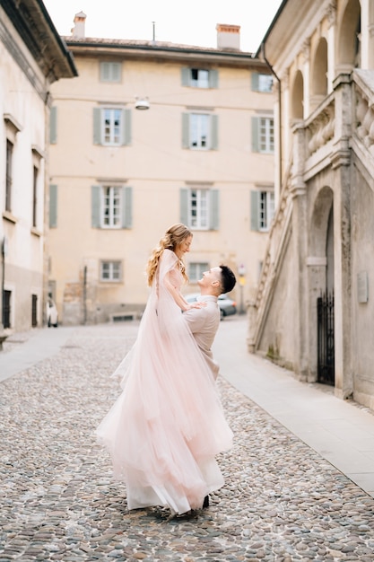 Groom holds bride in his arms while standing on the paving stones near the old building in bergamo