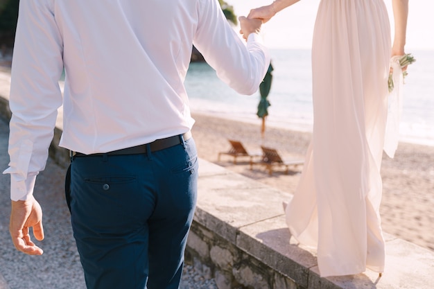 Groom holds the hand of bride with a bouquet walking along the seashore