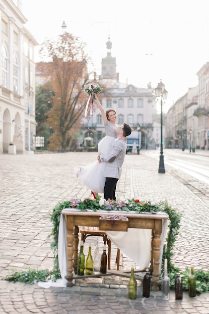 The groom holds his bride in his arms and stands against the background of old buildings in the city.