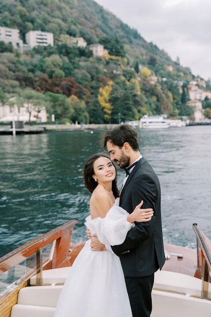 Groom hugs bride around the waist looking at her standing on a yacht sailing on the lake como italy