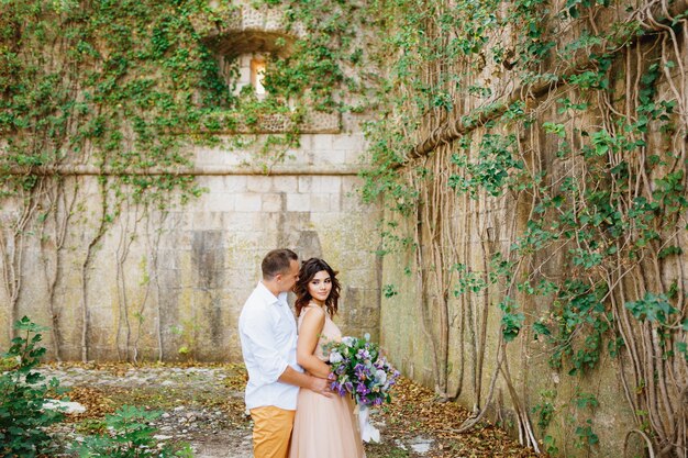 Groom hugs bride in a beautiful pastel dress with bouquet of flowers against the scene of a