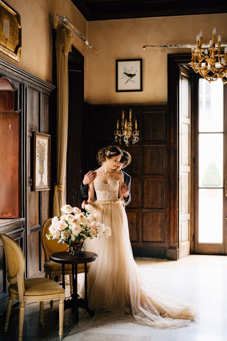 Photo groom hugs bride from behind near the table with a bouquet of roses in an old villa