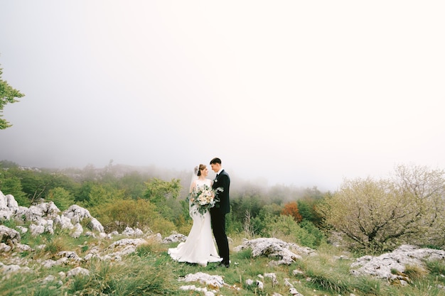Жених обнимает невесту в длинном платье с фатой и букетом цветов на фоне камней и
