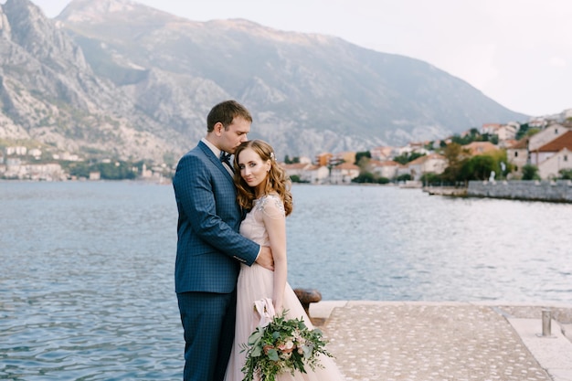 Groom hugs bride on the pier against the backdrop of mountains and the town on the coast