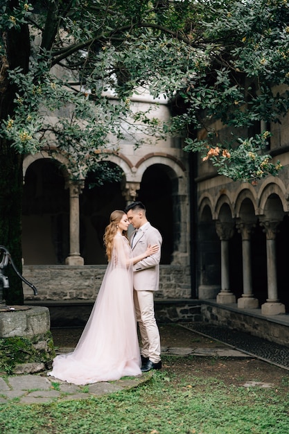 Groom hugs bride standing in a green garden against an old villa with columns