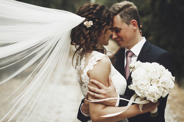 Photo groom hugs bride tender while wind blows her veil somewhere in tuscany, italy