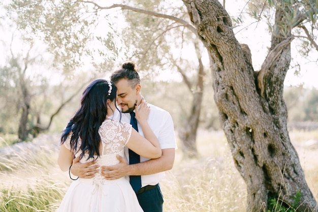 Groom hugs bride while standing by the tree in the park