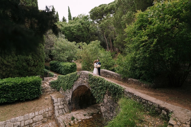 Groom hugs bride in a white dress with a bouquet of flowers standing on a stone bridge in a green