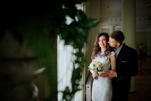 Groom hugs from behind pretty bride standing in the hall