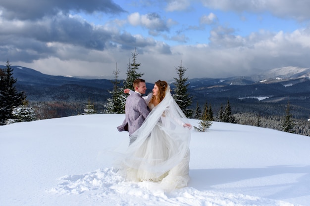 新郎は雪をかぶった山々を背景に彼の花嫁を抱きしめます。冬の結婚式の写真。