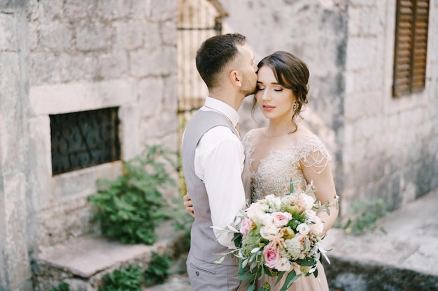 Groom kisses bride forehead near the stone building portrait