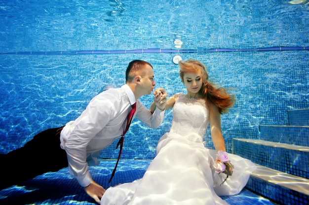 The groom kisses the hand of the charming bride underwater on a sunny day