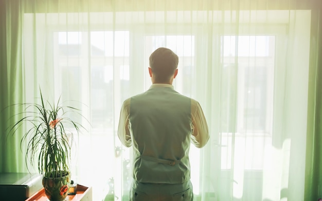 Жених в светло-сером костюме стоит у окна Мужчина смотрит перед собой