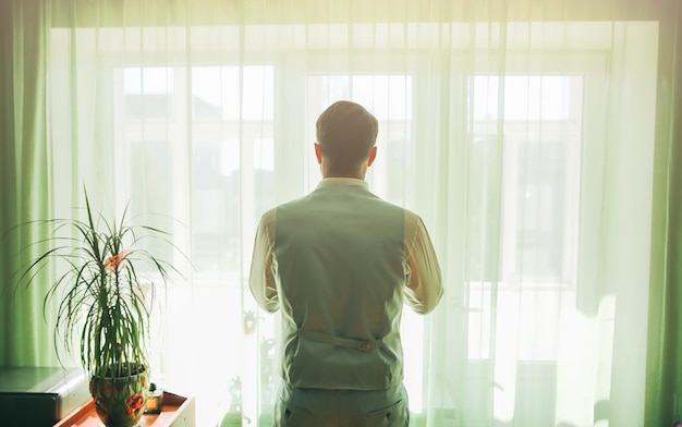 Groom in light grey suit is standing near the window Man looking in front of him