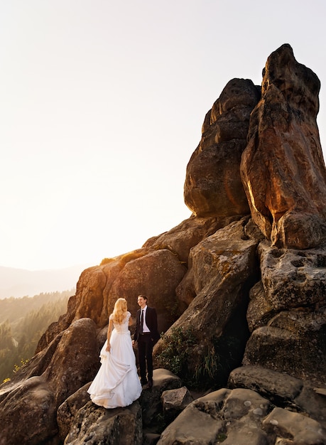 Groom looks at his wife while standing on the high enormous rock