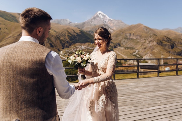 Groom in modern suit and bride in charming pink dress pose on the terasse with great mountain view in Georgia 