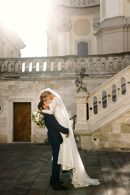 Groom raises bride up and she hugs him before white stairs to old church