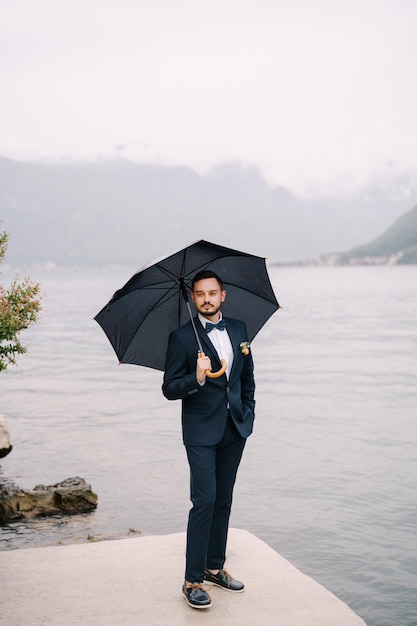 Photo groom stands under an umbrella on the pier by the water