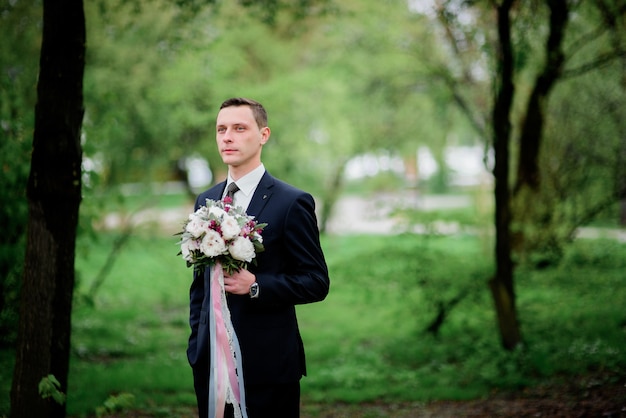 Groom stands with wedding bouquet in the forest