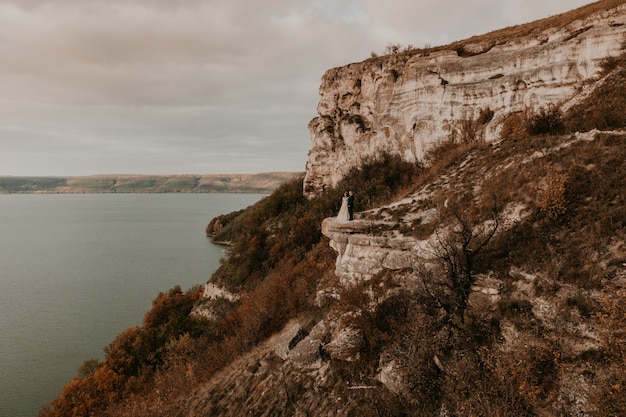 Groom in suit and the bride in a dress stand on a stone cliff above the river