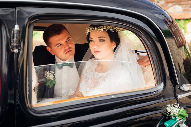 The groom in suit looks at a beautiful bride in a wedding dress and they are sitting in a car