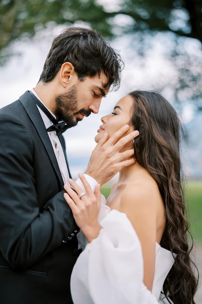 Photo groom touches bride face with his hand while standing in a green park