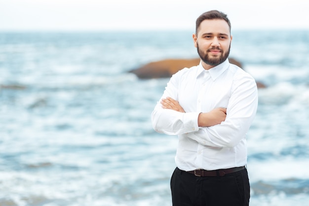 The groom in a white shirt and black pants stands on the beach