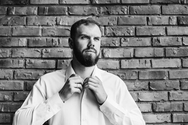 The groom in a white shirt fastens buttons on the background of a brick wall on black and white photography