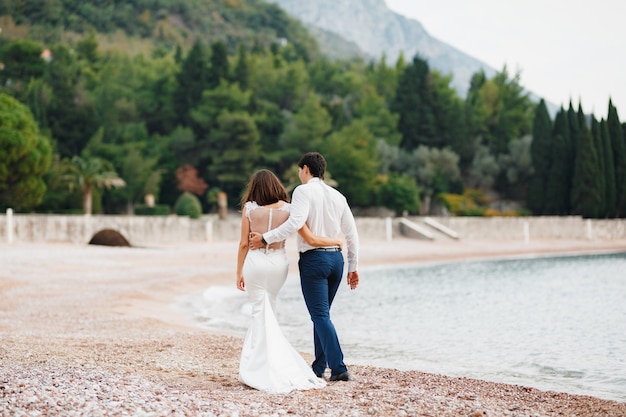 Жених в белой рубашке обнимает невесту в белом кружевном платье молодожены гуляют по пляжу спиной